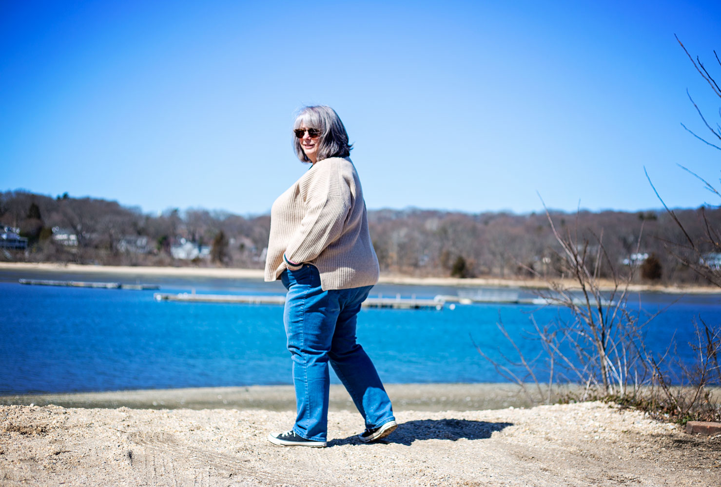 bettye in a beige sweater standing on a beach by the harbor with boats