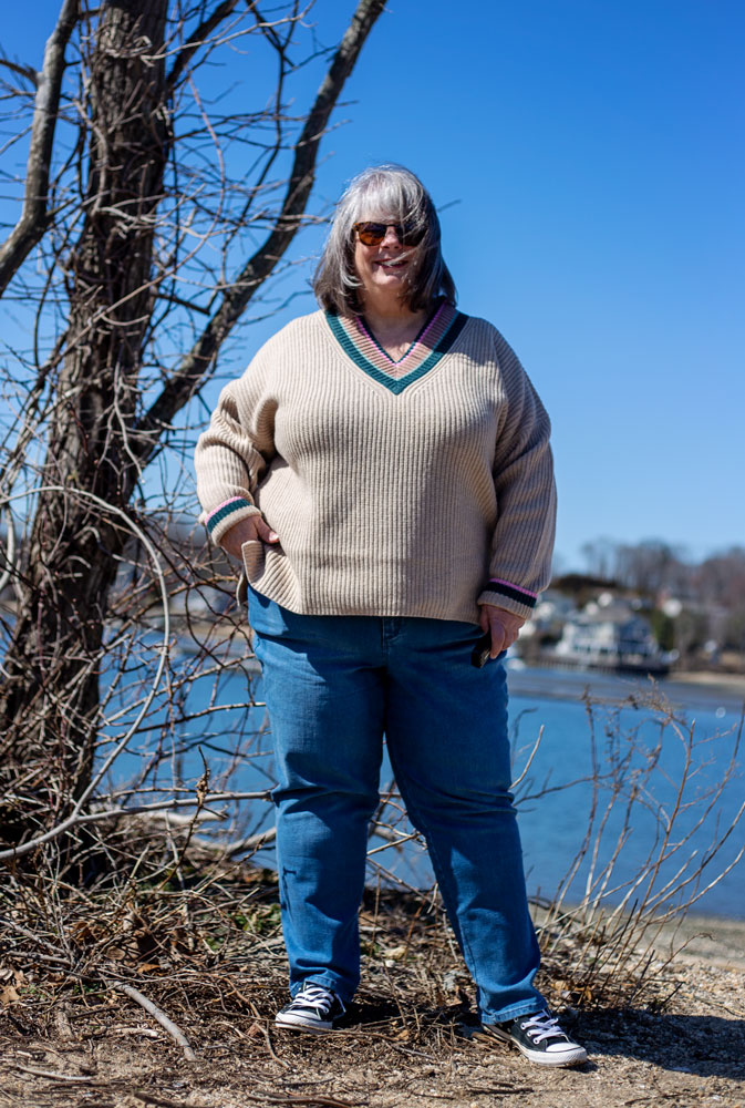 bettye standing next to a tree wearing a beige sweater and blue jeans in front of the harbor